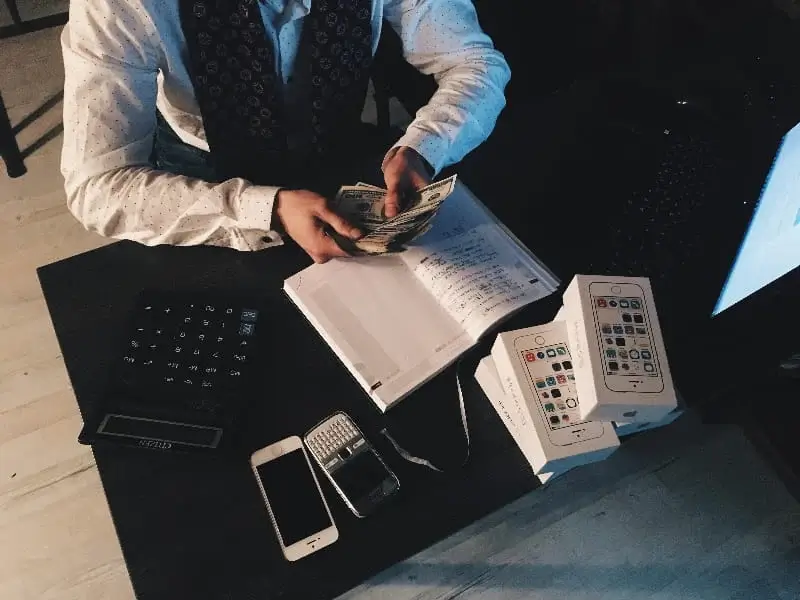 A person sitting at a table with a laptop and cell phone, counting money.