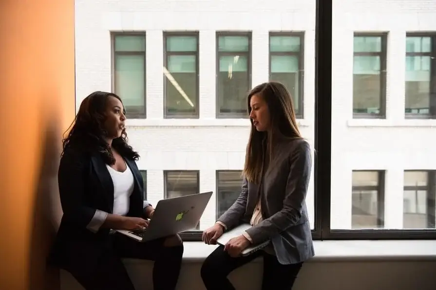 Two women engaged in a conversation while sitting in an office, using a laptop for communication.
